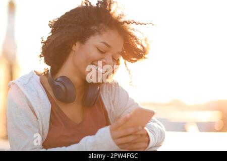 Outdoor portrait of happy cheerful african american woman in headphones using smartphone and smiling happily while standing at rooftop in morning Stock Photo
