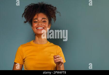Beautiful happy dark skinned african american ethnicity woman in yellow tshirt raising hand in greeting while looking in camera with pleasant smile Stock Photo