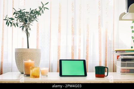 Digital tablet with green screen on wooden table with houseplant, candles, cup of tea and office supplies, cozy remote workplace with touch device. Stock Photo