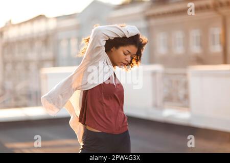 Full-length of young carefree african american woman jumping at rooftop in morning sunlight with cityscape on background, happy mixed-race female in Stock Photo