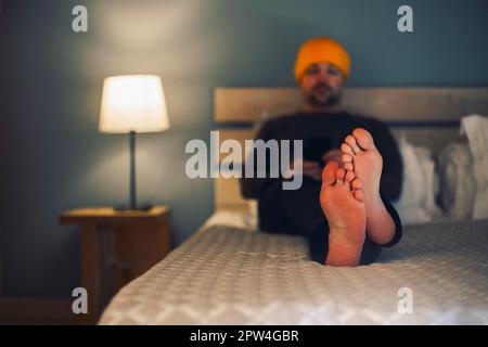 Portrait of mature handsome businessman sitting in yellow hat in hotel in rustic style with mobile phone. Man in formal wear sitting on bed using Stock Photo