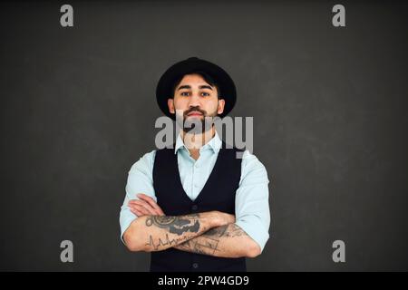 Studio portrait of handsome ethnic male with crossed tattooed arms, dressed in a blue shirt and black hat. Young stylish man on dark background Stock Photo
