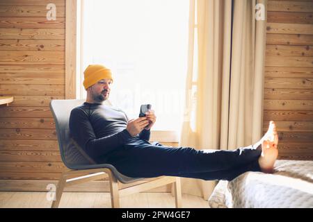 Portrait of mature handsome businessman sitting in yellow hat in hotel in rustic style with mobile phone. Man in formal wear sitting on bed using Stock Photo