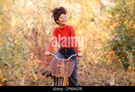 Young happy smiling african american woman riding bicycle in autumn forest, active positive mixed-race female in casual clothes smiling at camera Stock Photo