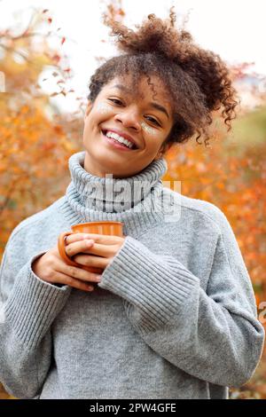 Young happy smiling mixed-race woman with coffee cup in autumn nature, pleased african american female with curly hair in knitted sweater looking Stock Photo