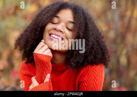 Young happy overjoyed african american woman with curly hair keeping eyes closed laughing and having fun while standing in autumn forest, enjoying Stock Photo