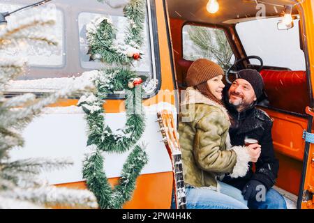 Happy middle-aged romantic couple in love sitting in minivan decorated with coniferous branches, drinking hot tea. Man and woman in warm clothes Stock Photo