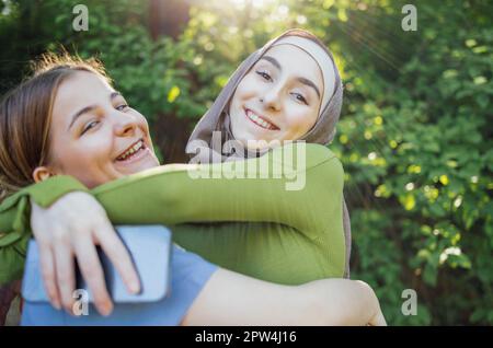 Strong female friendship. Happy two teen girls best friends holding hands and hugging while standing in front of park. Multiethnical friends Stock Photo