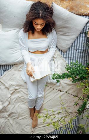Pretty romantic african american girl, reading book sitting on windowsill, suraunded with flowers. Beautiful Afro woman happily reading a novel in Stock Photo
