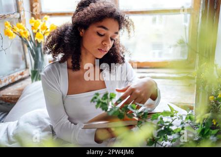 Pretty romantic african american girl, reading book sitting on windowsill, suraunded with flowers. Beautiful Afro woman happily reading a novel in Stock Photo