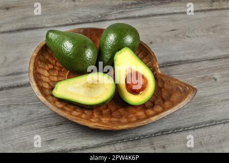 Three avocados, one of them cut to half, seed visible, in carved wooden bowl. Stock Photo
