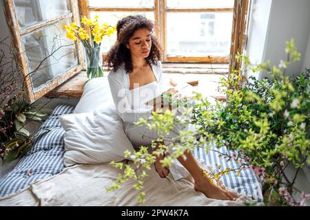 Pretty romantic african american girl, reading book sitting on windowsill, suraunded with flowers. Beautiful Afro woman happily reading a novel in Stock Photo