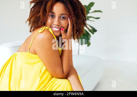 Beautiful curvy oversize young afto american woman in a yellow dress sitting on a sofa at home. Body positive stylish smiling female have relax time. Stock Photo