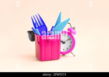 Alarm clock and plastic cutlery in trash bin on peach background. Time to say no to plastic cutlery. Plastic Pollution and Environmental Protection Co Stock Photo