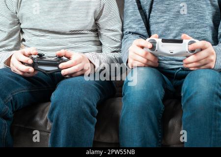 Male hands hold modified plastic joysticks. Friends play videogames sitting on couch. Two people hold wireless game console. Press buttons on controll Stock Photo