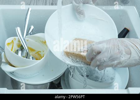 Human hands in protective domestic gloves wash white plate with sponge under tap water. Dish washing, tableware after meal, food leftovers. Pile of ki Stock Photo