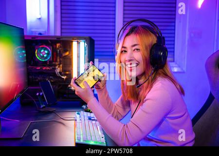 Gamer playing online game application on mobile phone wear gaming headphones looking to camera, Smiling young woman live stream she play video game vi Stock Photo