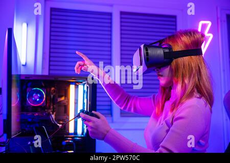 Gamer in VR headset glasses exploring metaverse plays online video game touching something on air with neon lights, Excited woman playing watching vid Stock Photo
