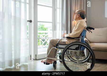 Asian senior man disabled sitting alone in wheelchair looking through window at hospital, lonely elder thoughtful sad old man look outside windows in Stock Photo