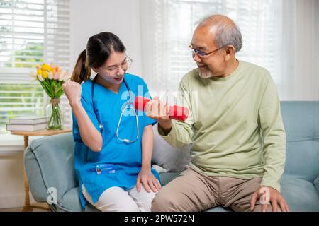 Young nurse physiotherapist helping old man in lifting dumbell at home, Asia nurse support elderly retirement sitting on sofa using red dumbbell worko Stock Photo