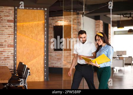 Two young focused coworkers, woman and man, looking through documents from folder walking in office. Business partners discussing financial graphs Stock Photo