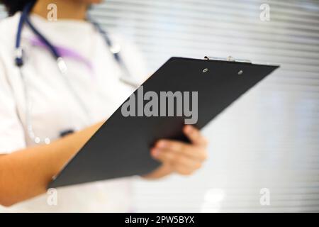 Ethnic female medic in mask and uniform filling medical report on clipboard in modern room in clinic. Healthcare concept Stock Photo
