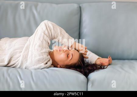 Portrait of relaxed positive young brunette woman lying down on soft blue sofa with eyes closed, pretty smiling while relaxing in cozy home Stock Photo