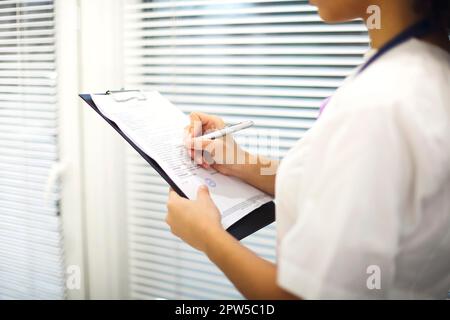 Ethnic female medic in mask and uniform filling medical report on clipboard in modern room in clinic. Healthcare concept Stock Photo