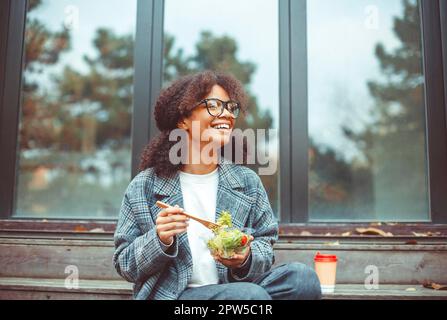 Happy african american woman office worker eating salad and smiling at camera while sitting on bench in park outdoors, selective focus. Positive Stock Photo