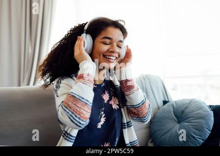 African millennial woman in orange sweater wearing white wireless headphones, listening music, resting and lying on couch in living room. Girl spend Stock Photo