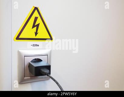 High-voltage sign on the white wall. A plug with a cable is stuck in an electrical outlet. Charging your phone or smartphone. The danger of electric s Stock Photo