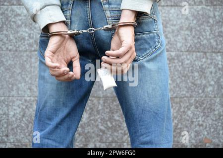 Arrested drug dealer in handcuffs with small heroin drug package on dark wall background. Illegal drug trafficking concept Stock Photo