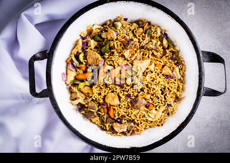 Japanese yakisoba with stir-fried noodles in metallic white pan on table with white tablecloth and black background. Top view. Stock Photo
