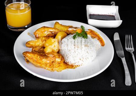 homemade chicken stew with tomatoes, onions and potatoes on a black fabric table and plate with rice, beans and orange juice. Stock Photo