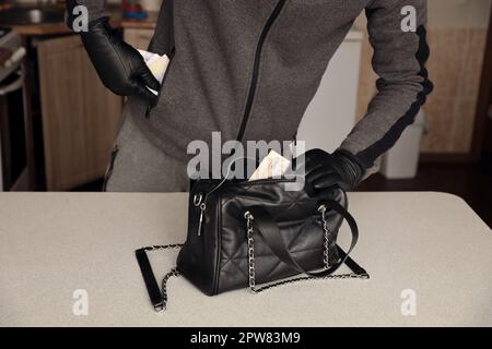 Robber in black outfit and gloves see in opened stolen women bag. The thief takes out the ukrainian money from a womans handbag in kitchen interior Stock Photo