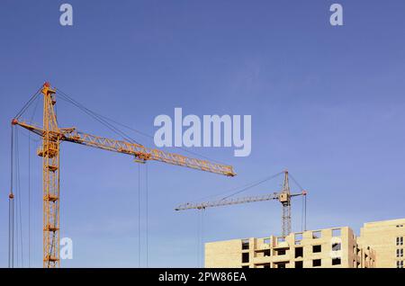 Working tall cranes inside place for with tall buildings under construction against a clear blue sky. Crane and building working progress with copyspa Stock Photo