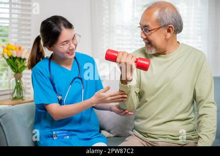 Asian nurse physiotherapist helping senior man in lifting dumbell at retirement home, Young nurse take care support training elderly sitting on sofa u Stock Photo