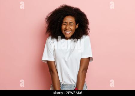 A portrait shot dark skinned curly haired shy and smiling woman in white t-shirt next to pink wall. High quality photo Stock Photo