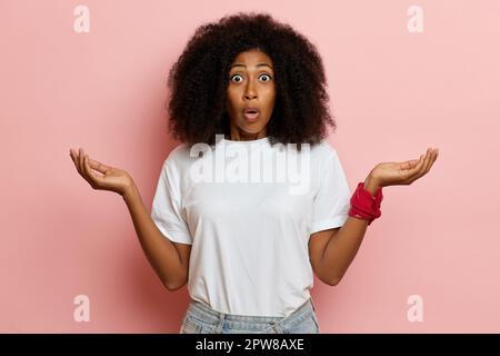 African American young woman, looks to the camera with wide opened eyes, puts hands a side in a goofy surprise. High quality photo Stock Photo