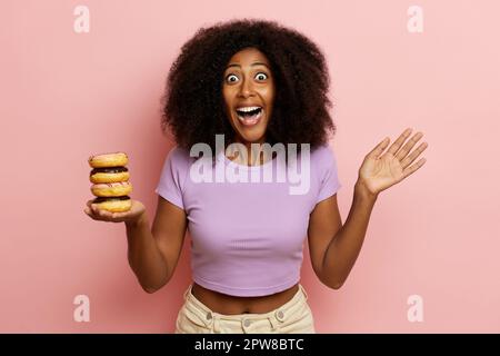 Photo of positive curly haired girl with surprised expression, raises palm, holds tasty doughnuts in one hand and looks directly to the camera with wi Stock Photo