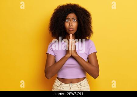Cute hopeful dark haired female model keeps palms pressed together under chin, prays for good luck, poses over yellow background. High quality photo Stock Photo