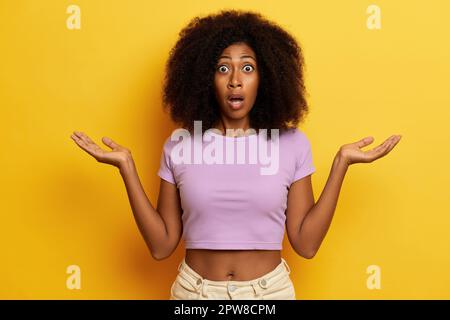Shocked curly haired woman keeps mouth opened with hands aside in hesitant gesture, isolated over yellow background. High quality photo Stock Photo
