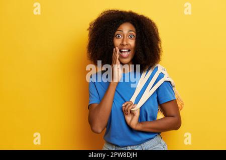 Amassed dark skinned curly haired woman, stands next to the yellow wall with wide opened eyes. Stock Photo