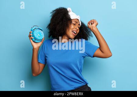 Curly haired African woman dancing and singing with alarm clock and sleeping mask on the head, she is happy and positive. Stock Photo