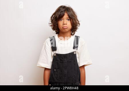 Studio shot of young disappointed teen guy with pouts lips, looks offended, wears denim overall, poses over white wall. High quality photo Stock Photo