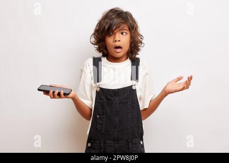 Portrait of confused brunette young guy, holds mobile phone, hesitates to make a decision, wears denim overalls, isolated over white wall. High qualit Stock Photo