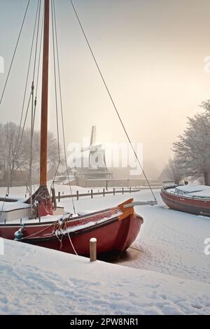 The Netherlands, Sloten, Traditional sailing cargo ship in frozen canal. Background windmill. Snow, winter. Stock Photo