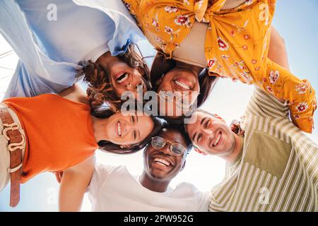 Low angle view of a group of five multiracial teenagers smiling and looking down at camera outdoors. Young student people of diverse ethnic cultures embracing while standing in a circle,together. High quality photo Stock Photo