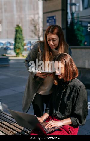 Two young, attractive businesswomen working together out in the city. Brunette businesswoman sitting on a bench and working on the lap top while her b Stock Photo