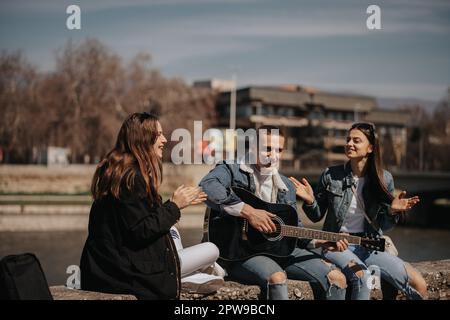 Handsome boy is playing guitar while sitting with his two girlfriends that are applauding him near the river Stock Photo